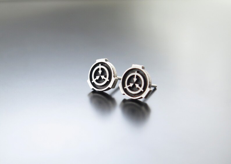 May include: A pair of silver stud earrings featuring a black and white symbol with three arrows pointing in different directions. The symbol is surrounded by a silver ring.