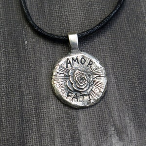 May include: Silver pendant necklace with a black cord. The pendant is round and features a rose with the words "AMOR FATI" inscribed around it.