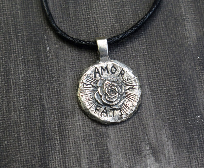 May include: Silver pendant necklace with a black cord. The pendant is round and features a rose with the words "AMOR FATI" inscribed around it.