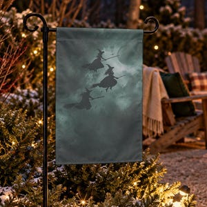 Könnte beinhalten: Eine rechteckige Gartenflagge mit einem dunklen, stimmungsvollen und bewölkten Himmelshintergrund, auf dem drei schwarze Silhouetten von Hexen auf Besenstielen zu sehen sind. Die Flagge ist an einem schwarzen Metallständer befestigt und detailliert.