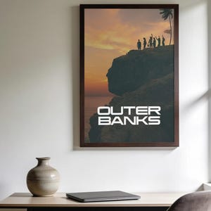 May include: A framed wall poster featuring a sunset landscape with silhouettes of people standing on a cliff edge near a palm tree. The text OUTER BANKS is printed in white capital letters at the bottom of the image within the dark frame.