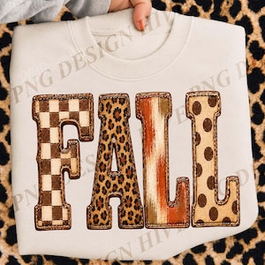 Op de afbeelding: Een crèmekleurig sweatshirt met ronde hals en het woord FALL in grote blokletters. Elke letter heeft een eigen patroon: de F is bruin-wit geblokt, de A heeft een luipaardprint, de L is oranje-bruin gestreept en de L is voorzien van stippen.