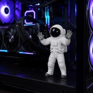 Puede incluir: Una figura de astronauta blanca con visera negra se encuentra con los brazos levantados dentro de una carcasa de ordenador. La carcasa presenta componentes negros, ventiladores y una vibrante iluminación LED morada y azul.