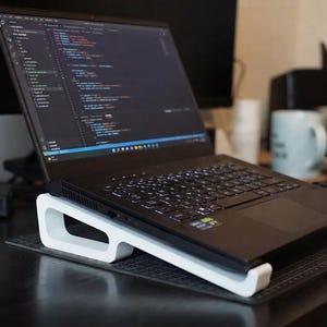 Może przedstawiać: Czarny laptop umieszczony jest na białej, zakrzywionej podstawce o otwartej konstrukcji. Ekran wyświetla linie kodu w edytorze z ciemnym motywem. Klawiatura jest czarna z podświetlanymi klawiszami, a urządzenie spoczywa na ciemnej, teksturowanej macie.