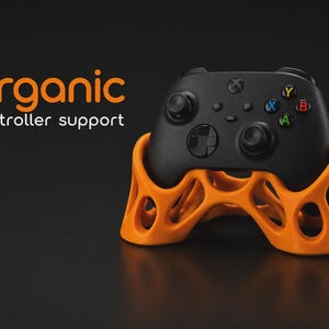 May include: A black video game controller resting on an orange, lattice-structured stand. The text "organic controller support" is printed in orange and white on the left side. The stand features a hollow, interconnected geometric design.