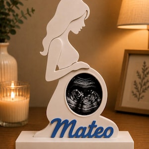 Può includere: Una sagoma bianca di una persona incinta in ginocchio su una base rettangolare. La figura presenta un ritaglio circolare nell'area dell'addome contenente un'immagine ecografica in bianco e nero. Il nome Mateo è scritto in lettere blu in corsivo.