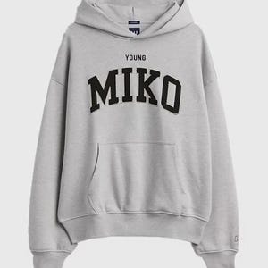 Pode incluir: Um moletom com capuz cinza-claro com mangas compridas, um bolso frontal tipo canguru e punhos e bainha canelados. O peito exibe o texto YOUNG MIKO em letras pretas, em negrito e arqueadas. Um pequeno logotipo da marca Gap é visível.