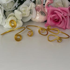 May include: Three gold-toned wire bracelets arranged in a row. Each bracelet features a distinct open-ended design with spiral or looped ends. One bracelet includes a small, pale pink stone accent secured within the wire wrapping.