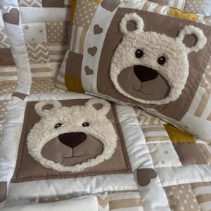 Könnte beinhalten: Eine Patchwork-Decke und ein passendes rechteckiges Kissen mit einer cremefarbenen, strukturierten Teddybären-Applikation. Das Stoffdesign besteht aus weißen, beigen und senfgelben Quadraten mit Stern-, Punkt- und Zickzackmustern, akzentuiert durch kleine braune Herzformen.