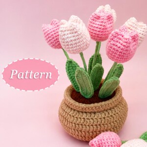 Peut inclure: Un arrangement de tulipes au crochet dans un pot beige avec des feuilles vertes et des fleurs roses et blanches. Le mot Pattern est imprimé en blanc dans un ovale rose. Deux tulipes au crochet supplémentaires sont posées devant le pot.