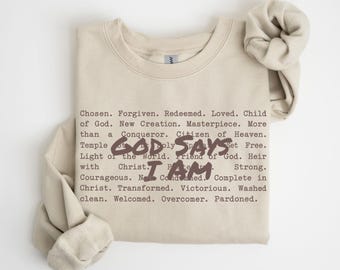 May include: A beige crewneck sweatshirt featuring dark brown text that reads "GOD SAYS I AM" in a large, bold font, surrounded by smaller, typewriter-style text listing various affirmations such as "Chosen," "Forgiven," and "Redeemed."