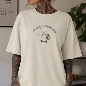 May include: A cream-coloured short-sleeve t-shirt featuring a graphic of a skeleton riding a scooter and carrying a basket of books. The text above the graphic reads "OFF TO TEACH SPOOKY MATH" in a black, arched, sans-serif font style.