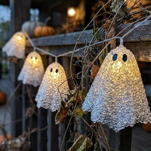 Pode incluir: Um cordão de luzes decorativas em forma de fantasma, com corpos translúcidos e texturizados feitos de pequenas contas transparentes. Cada fantasma possui dois olhos ovais escuros e é iluminado internamente por uma luz branca quente.