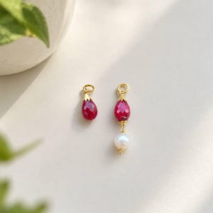 May include: Two gold-toned jewelry charms. One charm features a single faceted, teardrop-shaped deep pink gemstone. The second charm features a faceted, teardrop-shaped deep pink gemstone suspended above a single round white pearl.