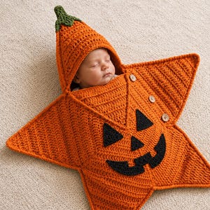 May include: A star-shaped orange crochet baby swaddle blanket featuring a jack-o'-lantern face with black eyes, nose, and mouth. The swaddle includes a pointed hood with a green stem top and three wooden buttons along the side edge.