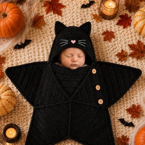 May include: A black crocheted star-shaped baby swaddle blanket featuring a hood with cat ears, white embroidered whiskers, and a small pink heart nose. The swaddle includes three light brown wooden buttons along one side to secure the garment.