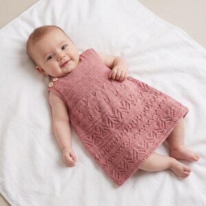 May include: A dusty rose knitted baby dress featuring a sleeveless design, a garter stitch bodice, and a detailed lace pattern on the skirt. The garment is secured at the shoulder with two small, light-colored wooden buttons, providing a simple closure.