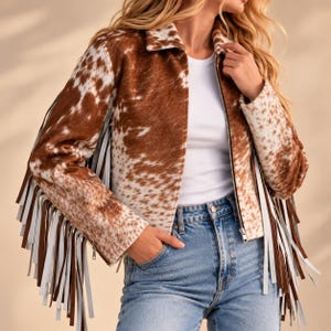 Puede incluir: Una chaqueta con estampado de vaca en marrón y blanco que presenta un cuello clásico, una cremallera frontal completa y mangas largas adornadas con tiras de flecos alternas en marrón y blanco que van desde los hombros hasta los puños.