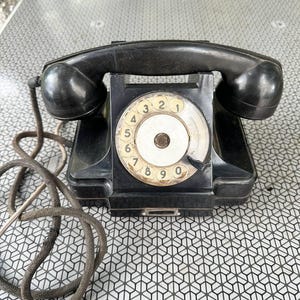Puede incluir: Un teléfono de disco negro vintage que presenta un dial circular con números del cero al nueve, un auricular a juego apoyado en la parte superior y un cable grueso cubierto de tela que se extiende desde el lado de la base rectangular.