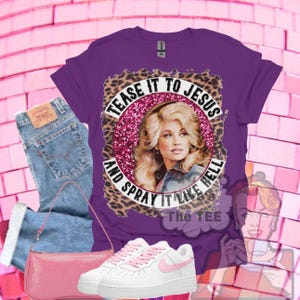 Könnte beinhalten: Ein violettes T-Shirt mit einer Grafik einer blonden Frau in einem pinken Glitzerkreis mit Leopardenmuster-Rand. Der Text lautet TEASE IT TO JESUS AND SPRAY IT LIKE HELL. Ebenfalls enthalten sind eine blaue Jeans, eine pinke Handtasche und weiße Sneaker.