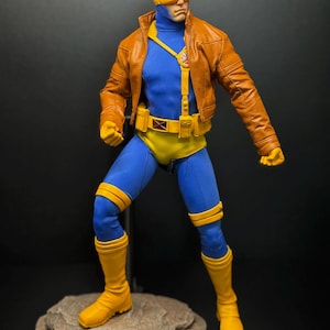 May include: A collectible action figure of a character wearing a brown leather-style jacket, a blue bodysuit, yellow boots, yellow gloves, and a yellow utility belt with a red X emblem. The figure wears a yellow visor over the eyes.