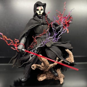May include: A posable action figure of a character wearing a black hooded robe and a skull-like mask. The figure holds a red lightsaber and is surrounded by translucent red and purple lightning effects, standing on a brown textured base.