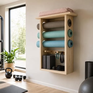 May include: A wall-mounted light wood storage rack featuring three horizontal shelves for rolled yoga mats, a top shelf holding a folded pink blanket, and lower pegs for hanging resistance bands, alongside two black foam blocks and accessories.