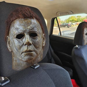 May include: A realistic Michael Myers mask with textured, weathered skin, deep-set black eye sockets, and brown synthetic hair attached to the top, mounted securely onto the black fabric headrest of a vehicle seat for display purposes.