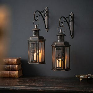 May include: Two wall-mounted lanterns with a dark bronze finish and clear glass panels. Each lantern features a decorative scrollwork bracket and a hinged door. Inside each lantern, a warm glowing candle is visible, providing soft illumination.