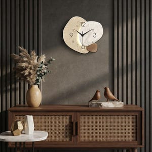 May include: A wall clock with an irregular, organic shape featuring a textured beige section, a smooth white marble-patterned section, and a small brown accent piece. The clock face has black numerals and thin black hands indicating the time.