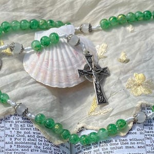 May include: A rosary featuring translucent green beads interspersed with smaller white beads, a silver-toned crucifix with intricate detailing, and a circular silver-toned medal depicting a religious figure, all arranged in a circular formation.