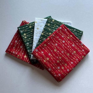 May include: A stack of four folded fabric squares in red, dark green, and white. Each square features a repeating pattern of vertical wavy lines interspersed with small gold and silver stars. The fabric pieces are layered to show the colours.