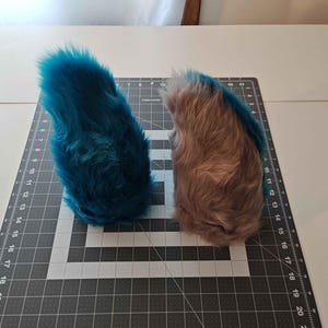 May include: Two faux fur tail accessories positioned on a black cutting mat with white grid lines. One tail is a vibrant teal blue, while the other is a light tan brown. Both items feature long, soft, synthetic fibers and a curved, tapered shape.