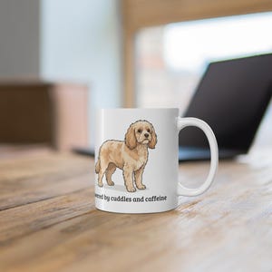 May include: A white ceramic mug featuring a cartoon illustration of a light brown curly-haired dog standing in profile. Below the dog, the text reads "powered by cuddles and caffeine" in a simple black sans-serif font. The mug has a handle.