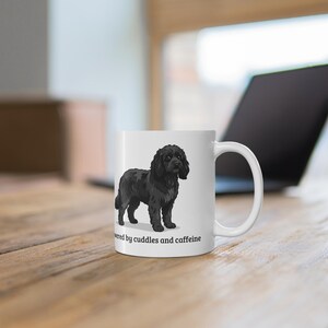 May include: A white ceramic mug featuring a black dog illustration and the text "powered by cuddles and caffeine" printed in black. The mug has a standard handle on the right side and a smooth, cylindrical body with a clean, white finish.