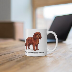 May include: A white ceramic mug featuring a brown spaniel dog illustration and the black text "powered by cuddles and caffeine" printed on the front. The mug has a standard cylindrical shape with a curved handle on the right side.
