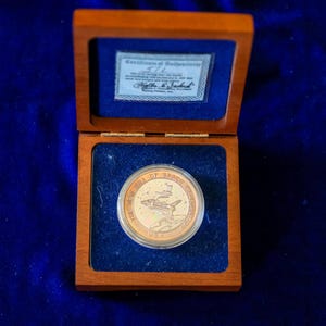 Puede incluir: Una moneda conmemorativa circular con el diseño de un transbordador espacial y el texto The New Era of Space Exploration U.S.A., dentro de una cápsula protectora transparente, en una caja de madera cuadrada con forro de terciopelo azul.