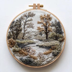 May include: A circular wooden embroidery hoop framing a landscape scene stitched with thread. The design features trees, bushes, and a central path in shades of beige, brown, gray, and white, creating a textured, detailed nature scene.