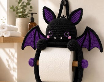 May include: A black and purple crocheted bat toilet paper holder featuring a round head with pointed ears, wide purple-accented wings, and two small white fangs. The bat holds a standard white roll of toilet paper on a wooden dowel rod.