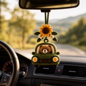 May include: A crocheted car ornament in dark green featuring a brown bear in the driver seat. Above the car is a yellow sunflower with white flowers and green leaves, all attached to a dark green crocheted hanging loop for a rearview mirror.