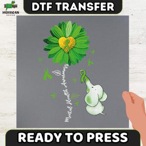 May include: A DTF transfer design on a gray background featuring a green daisy with a yellow heart center containing a green awareness ribbon. A small white elephant holds a green ribbon with its trunk. The text reads Mental Health Awareness.
