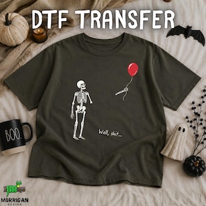May include: A dark olive green short-sleeve t-shirt featuring a white skeleton graphic. The skeleton is missing an arm, while a separate skeletal hand holds a red balloon. The text "Well, shit..." is printed in white below the skeleton.