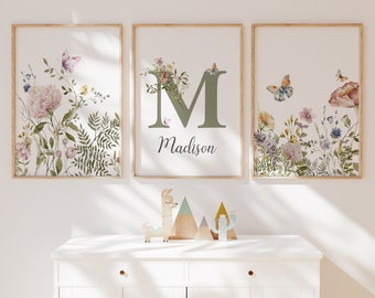 May include: Three framed prints with watercolor floral designs. The center print features the letter "M" in green with the name "Madison" written below. The prints are hung on a white wall.