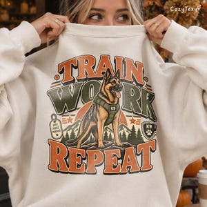Puede incluir: Una sudadera color crema con el gráfico de un pastor alemán con arnés táctico. El texto dice TRAIN WORK REPEAT con etiquetas de K9 y DOGS PEOPLE PURPOSE. El diseño incluye siluetas de montañas y bosques en tonos naranja y verde.