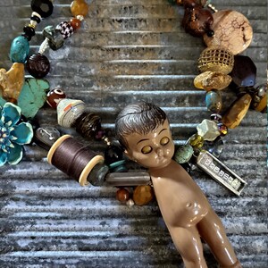 May include: A colorful beaded necklace with a small doll figure, a spool of thread, and a metal tag with the word "PRESSE" on it. The necklace is made with various materials, including beads, stones, and metal.