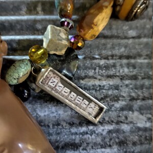 May include: A silver charm with the word "ZAPPER" spelled out in individual silver squares. The charm is attached to a necklace with various beads, including amber, turquoise, and glass.