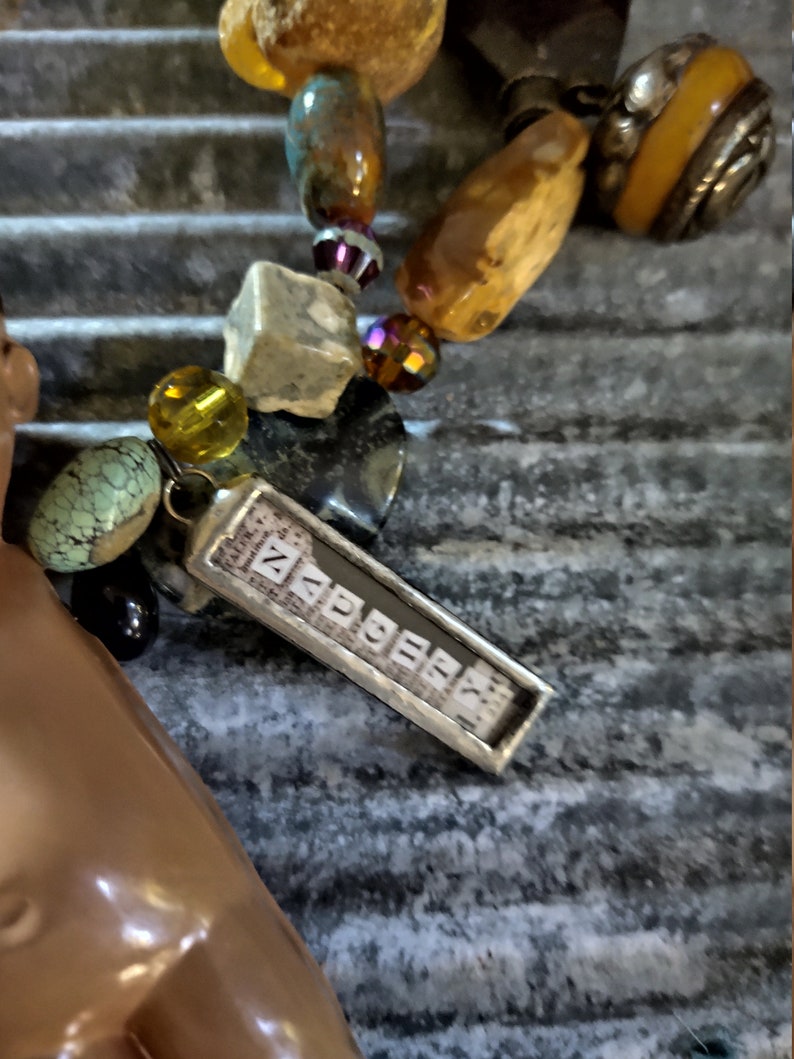 May include: A silver charm with the word "ZAPPER" spelled out in individual silver squares. The charm is attached to a necklace with various beads, including amber, turquoise, and glass.