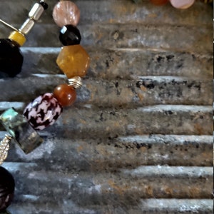 May include: A multi-colored beaded necklace with a silver clasp. The beads are made of various materials, including glass, stone, and metal. The necklace is strung on a brown cord.