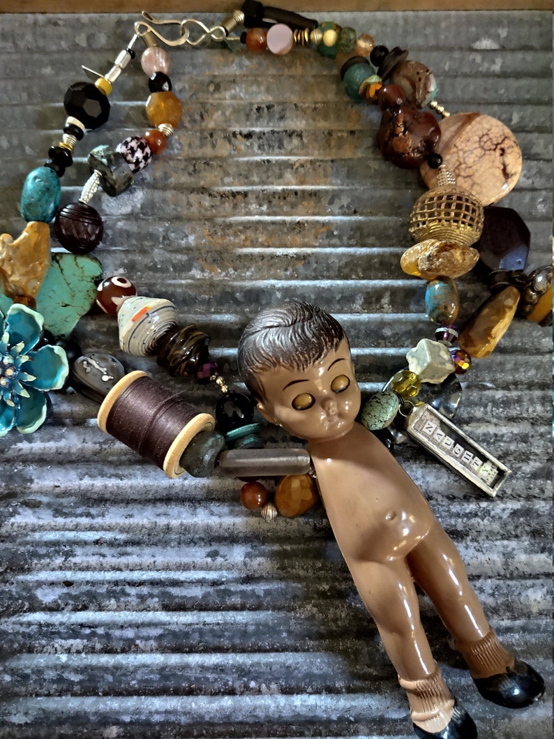 May include: A colorful beaded necklace with a small doll figure, a spool of thread, and a metal tag with the word "PRESSE" on it. The necklace is made with various materials, including beads, stones, and metal.