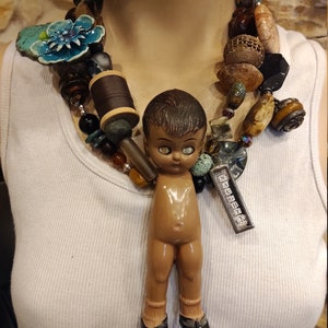May include: A multi-strand necklace with a variety of beads, including turquoise, brown, and gold. A small doll with brown hair and large eyes hangs from the center of the necklace. The doll is wearing black shoes and has a brown body.